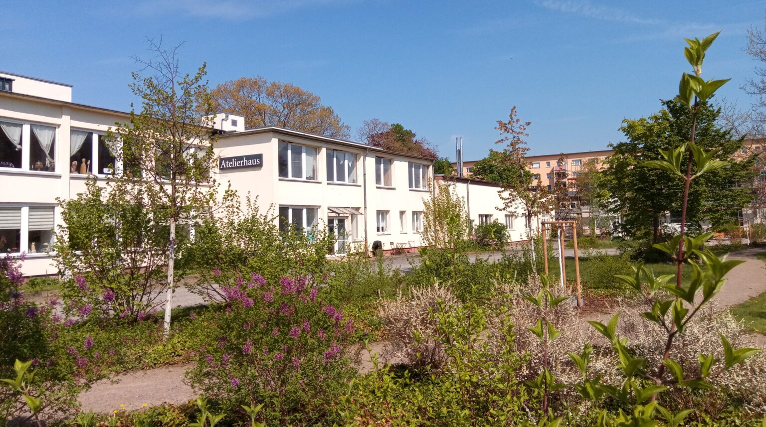 Ein Foto des Atelierhauses im Mai 2022. Bäume und Büsche mit weißen und pinkfarbenen Blüten im Vordergrund. Dahinter das Atelierhaus mit den Vereinsräumen in einem Blickwinkel von etwa 45 Grad.