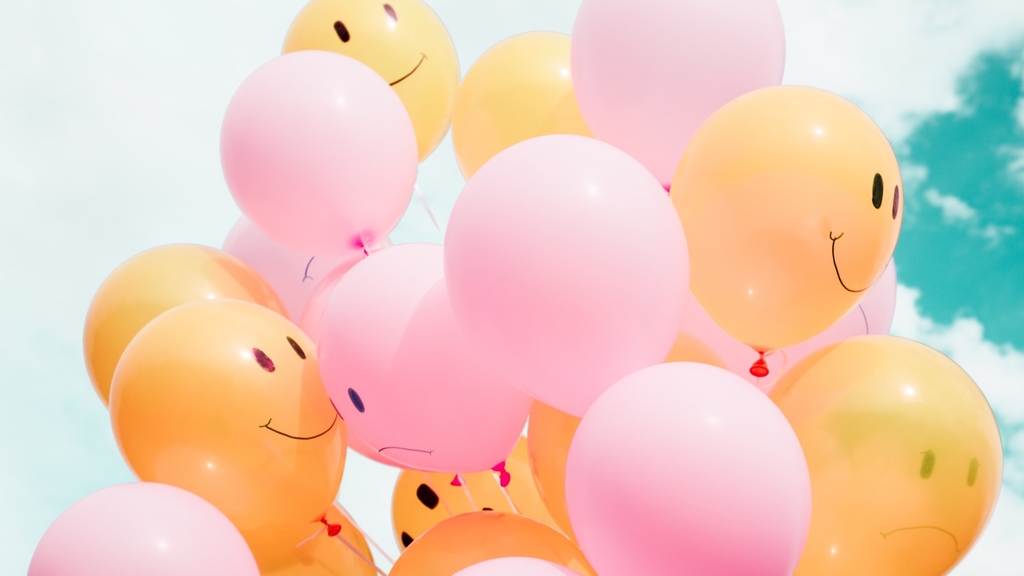 Foto eines "Straußes" von rosa und gelben Luftballons mit aufgemalten Augen und Mund, teils lachend, teils traurig vor einem türkisblauen Himmel mit weißen Wolken.