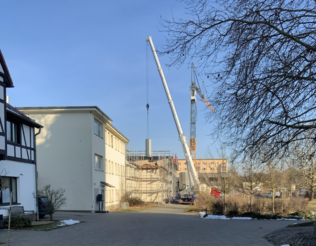 Blick den Weg hinab aufs Atelierhaus noch im gelben schäbigen Kleid, zum Teil mit Gerüst. Ein weißer Kran hebt irgendein Betonteil aufs Dach. Dahinter ein stationärer gelber Kran.