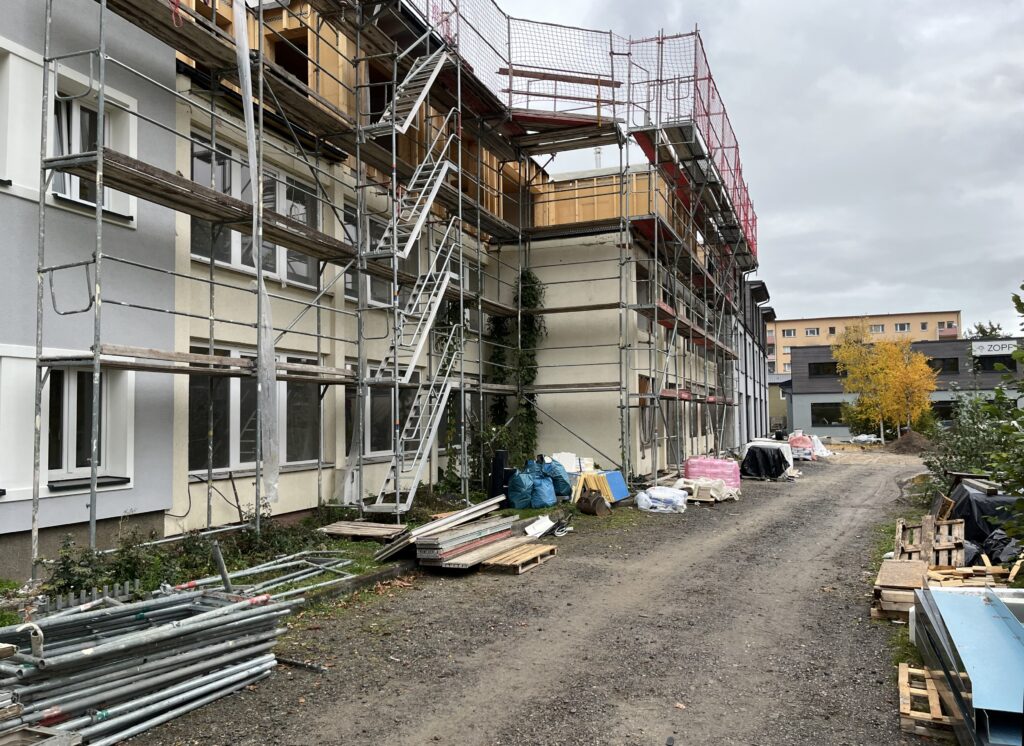 Am linken Rand das Atelierhaus schon im neuen grauen Anstrich übergehend in den mittleren Gebäudeteil noch im gelben, schäbigen Kleid mit Gerüst gefolgt vom hinteren Gebäudeteil auch schon im frischen Grau. Im Vordergrund liegen links des Weges Gerüstteile und rechts Palettenreste und Baumaterialien.