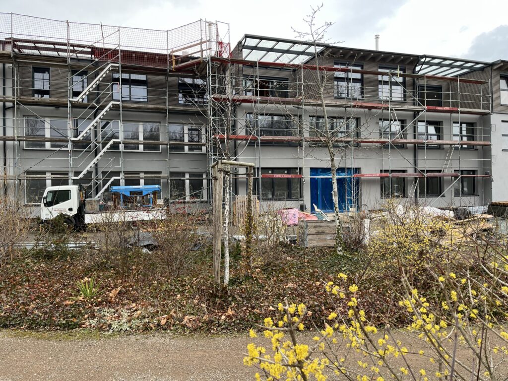 Im Hintergrund "unser" Mittelteil des Atelierhauses noch mit Gerüst aber schon mit zweitem OG und ganz in frischem Grau. In der linken Bildhälfte ein kleiner Laster, davor noch kahle Pflanzen. In den vierten Quadranten ragen etwas unscharf die leuchtend gelben Blüten des Zauberhaselstrauchs.