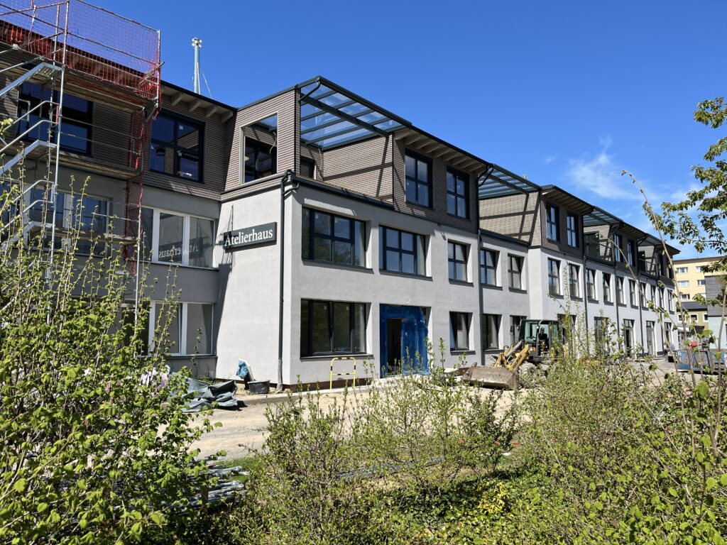 Über Büsche mit jungen grünen Blättern hinweg das Atelierhaus ohne Gerüst in frischem Grau. Der Radlader vor dem Eingang unseres Mittelteils und etwas Unordnung ringsherum zeugen noch vom laufenden Innenausbau.